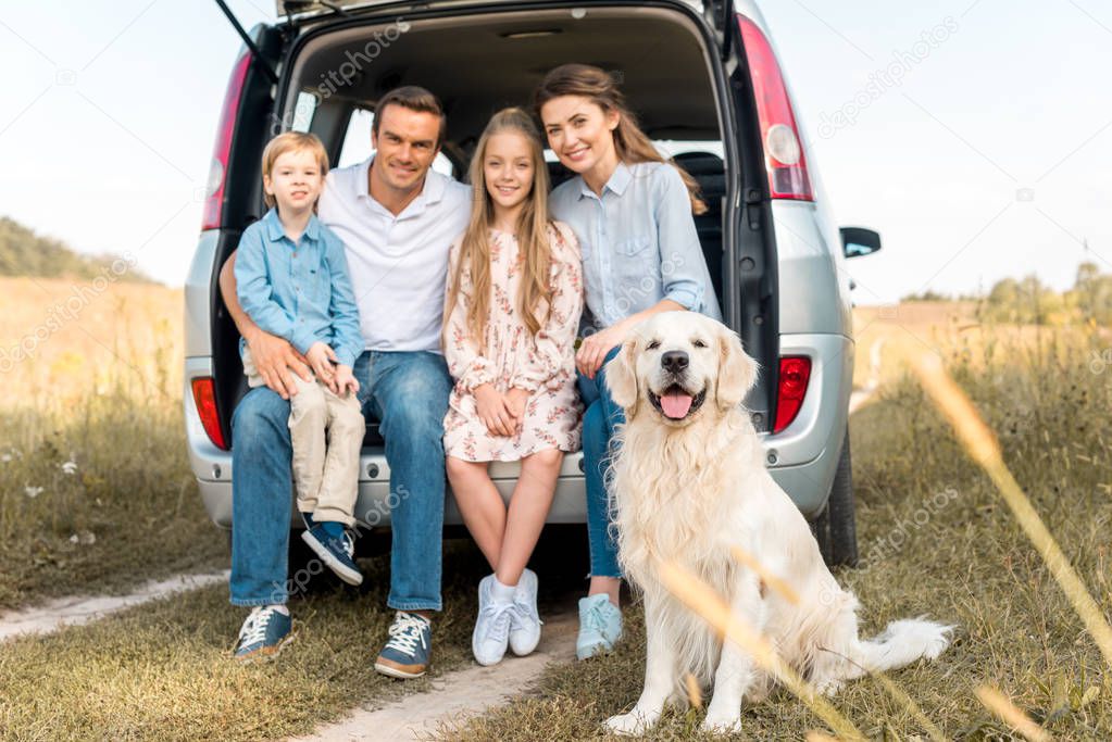 happy young family with retriever dog sitting in car trunk and looking at camera in field