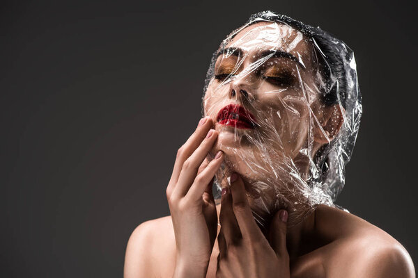 sensual woman with face wrapped in transparent cellophane with drops isolated on black 