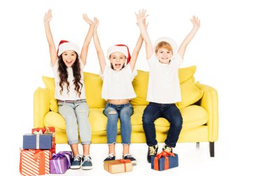 Noel Baba şapkaları kaldırdı elleriyle beyaz izole sarı koltukta oturan mutlu sevimli çocuklar