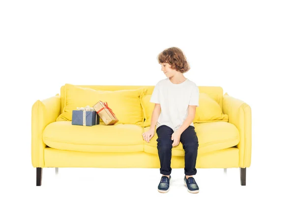 Adorable boy sitting on yellow sofa and looking at gift boxes isolated on white — Stock Image Adorable Boy Sitting Yellow Sofa Looking Gift Boxes Isolated White Stock Image