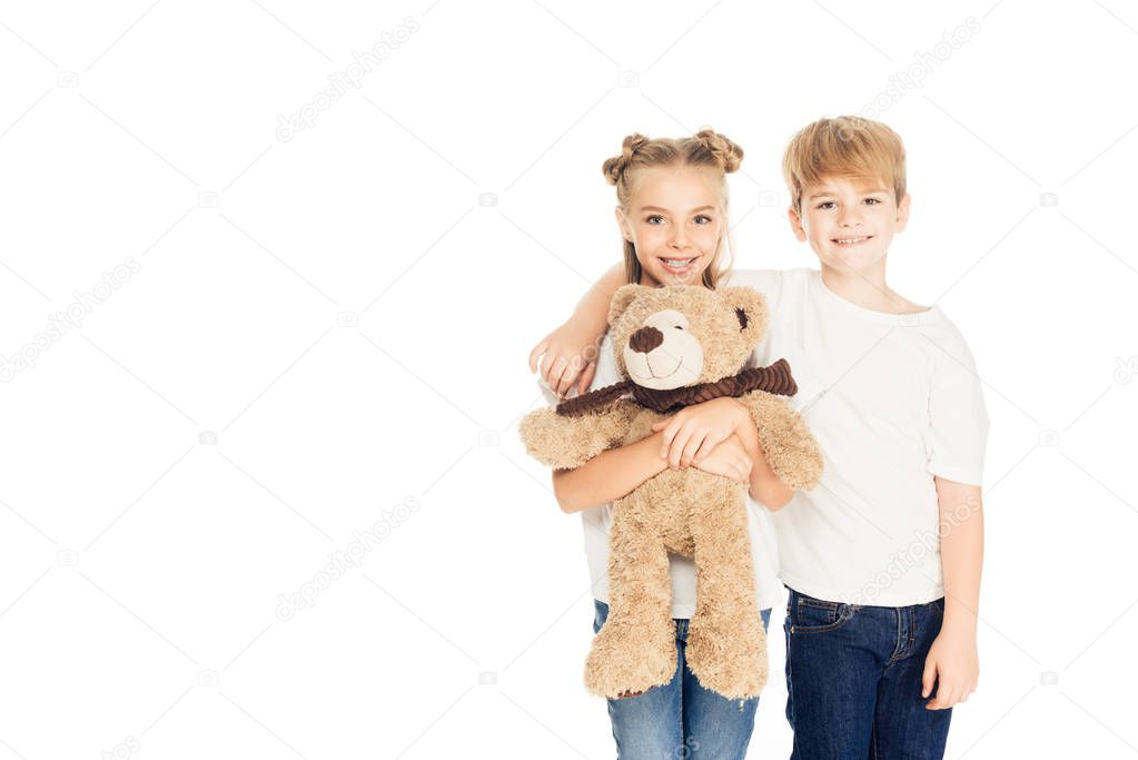 cheerful kids hugging, holding teddy bear and looking at camera isolated on white