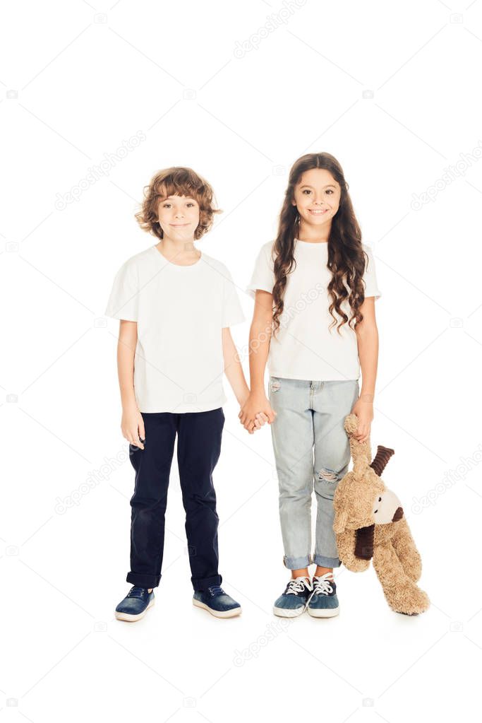 smiling adorable kids holding hands and teddy bear isolated on white, looking at camera