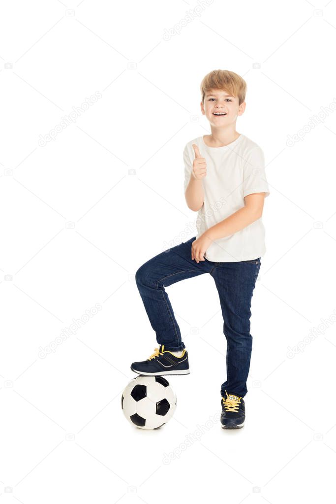 smiling adorable boy putting leg on football ball and showing thumb up isolated on white