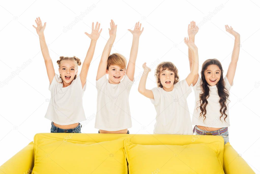 happy children standing behind yellow sofa with raised hands isolated on white