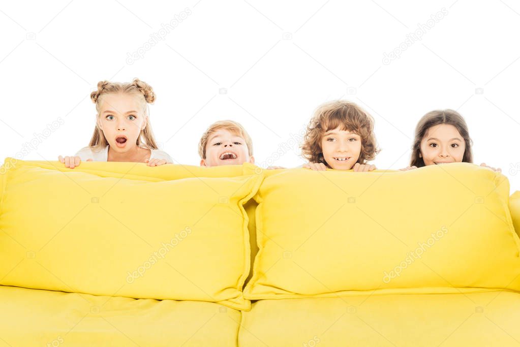 excited and smiling children looking out from yellow sofa and looking at camera isolated on white
