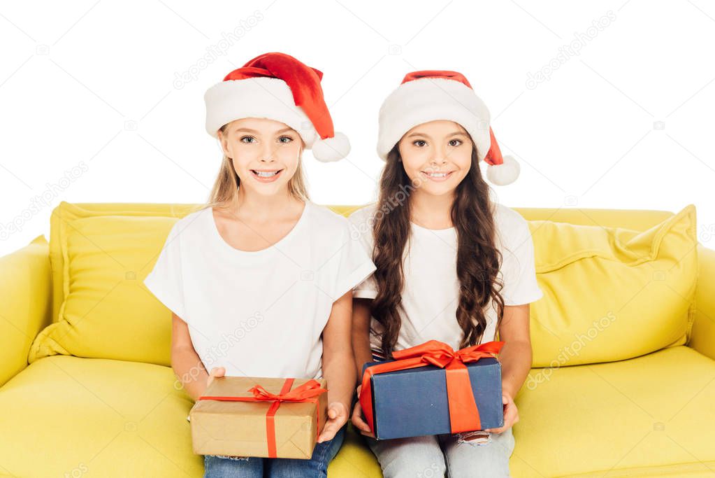 smiling adorable kids in santa hats sitting on yellow sofa with presents isolated on white