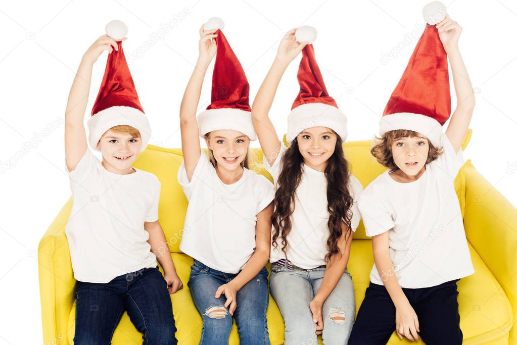 smiling adorable kids holding santa hats and sitting on yellow sofa isolated on white