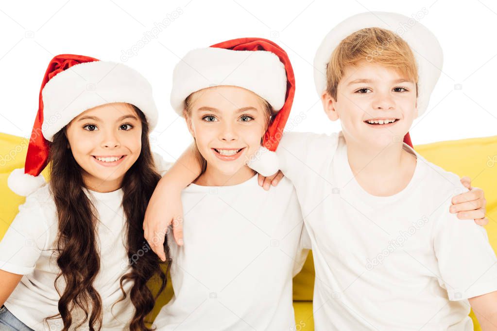 portrait of smiling adorable kids in santa hats sitting on yellow sofa and looking at camera isolated on white
