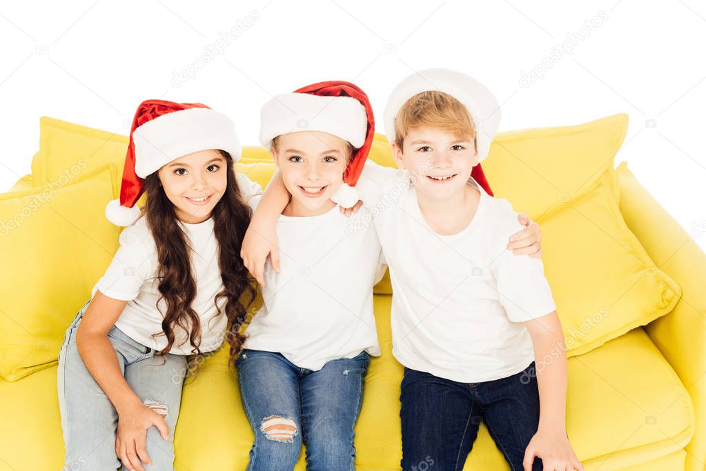 high angle view of smiling adorable kids in santa hats sitting on yellow sofa and looking at camera isolated on white