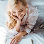 Vista de ángulo alto de hermosa chica rubia feliz en pijama acostado en la cama y libro de lectura