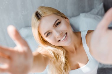 selective focus of happy blonde woman reaching hands and smiling at camera in bedroom clipart