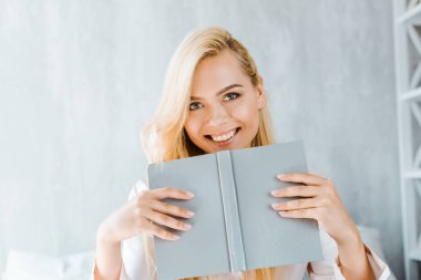 happy young woman holding book and smiling at camera in bedroom clipart
