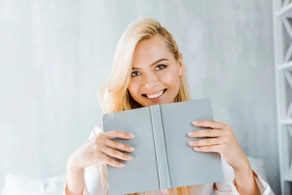stock image happy young woman holding book and smiling at camera in bedroom