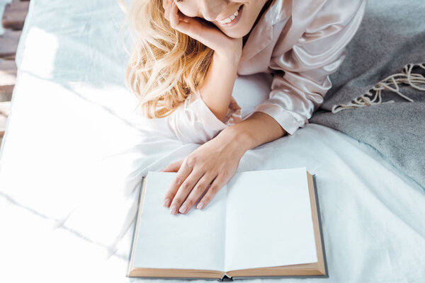 cropped shot of smiling young woman lying on bed and reading book