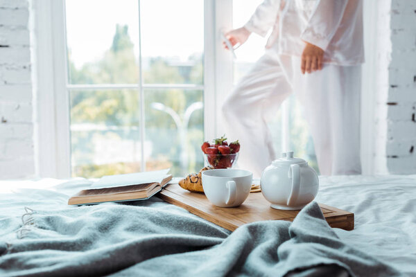 cropped image of woman opening window, breakfast and book on bed