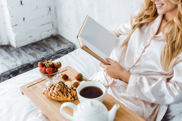 cropped image of attractive woman in pajamas reading book, breakfast on wooden tray in bed