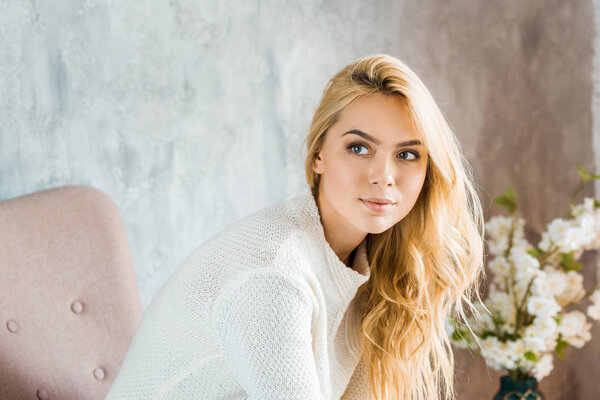 portrait of beautiful woman in sweater sitting on armchair in bedroom and looking away