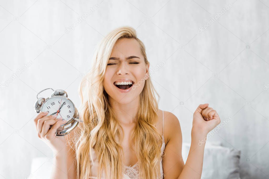 young blonde woman holding alarm clock and yawning in bedroom