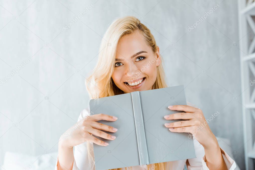 happy young woman holding book and smiling at camera in bedroom