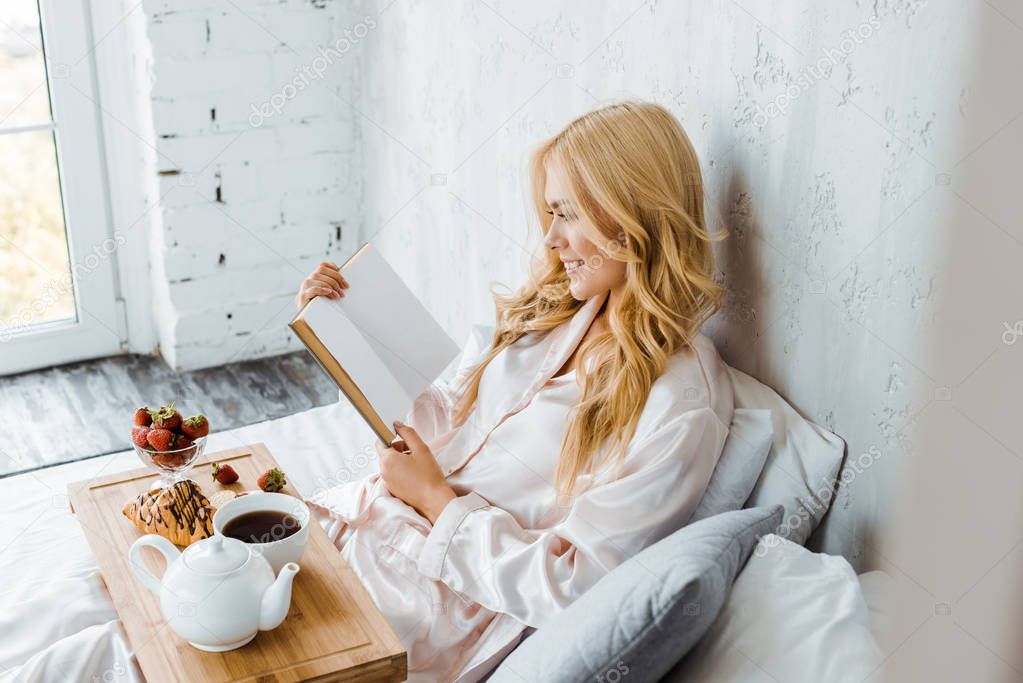 attractive woman in pajamas reading book, breakfast on wooden tray in bed