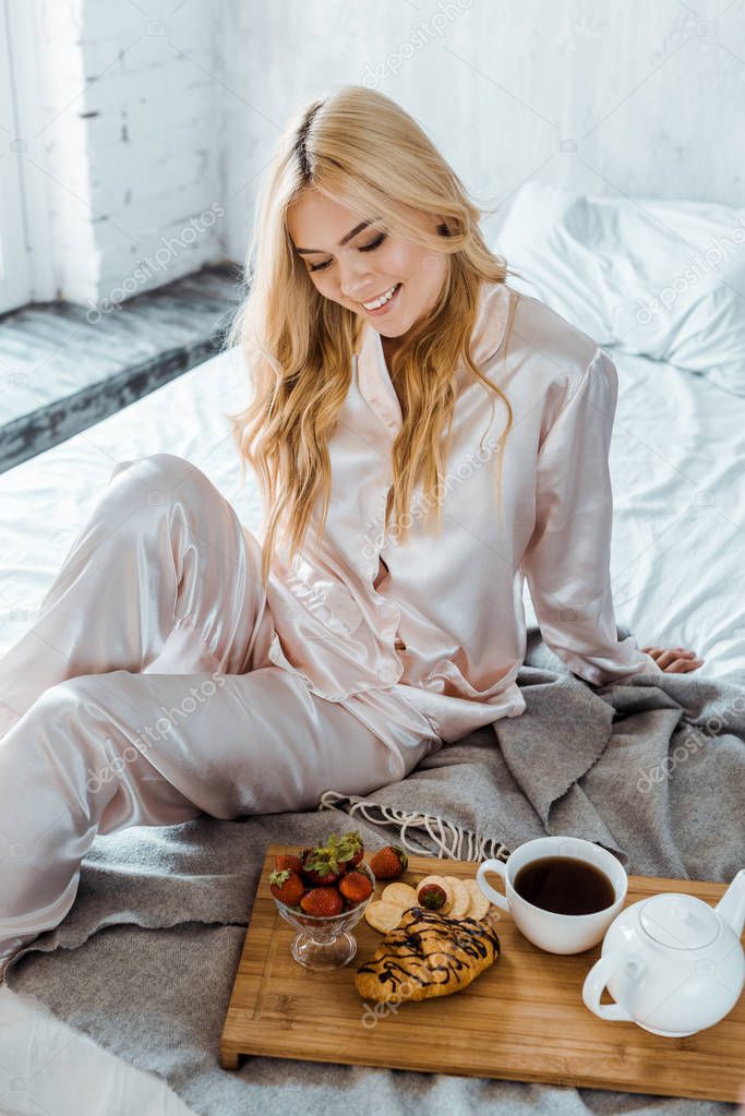 smiling attractive woman in pajamas looking at breakfast on wooden tray in bed