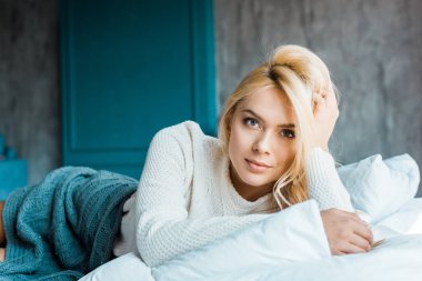 Yatak odasında yatakta battaniyenin altında yalan ve kameraya bakarak kazaklı çekici kadın