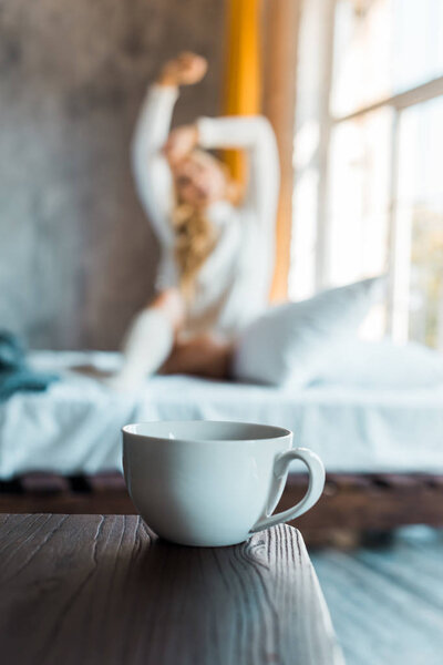 selective focus of attractive woman in sweater stretching in bedroom in morning, cup of coffee on tabletop