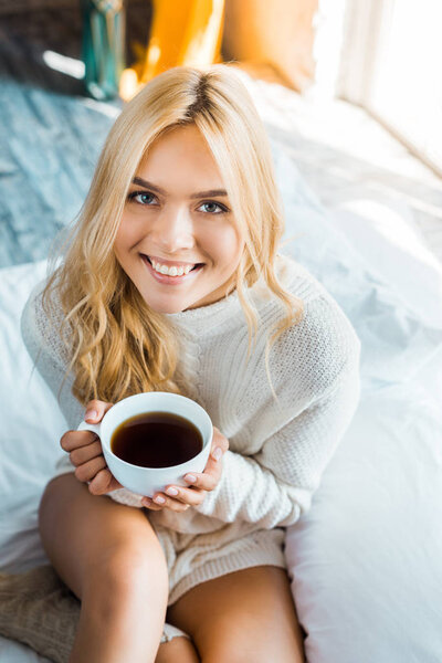 smiling attractive woman in sweater holding cup of coffee and looking at camera in bedroom in morning