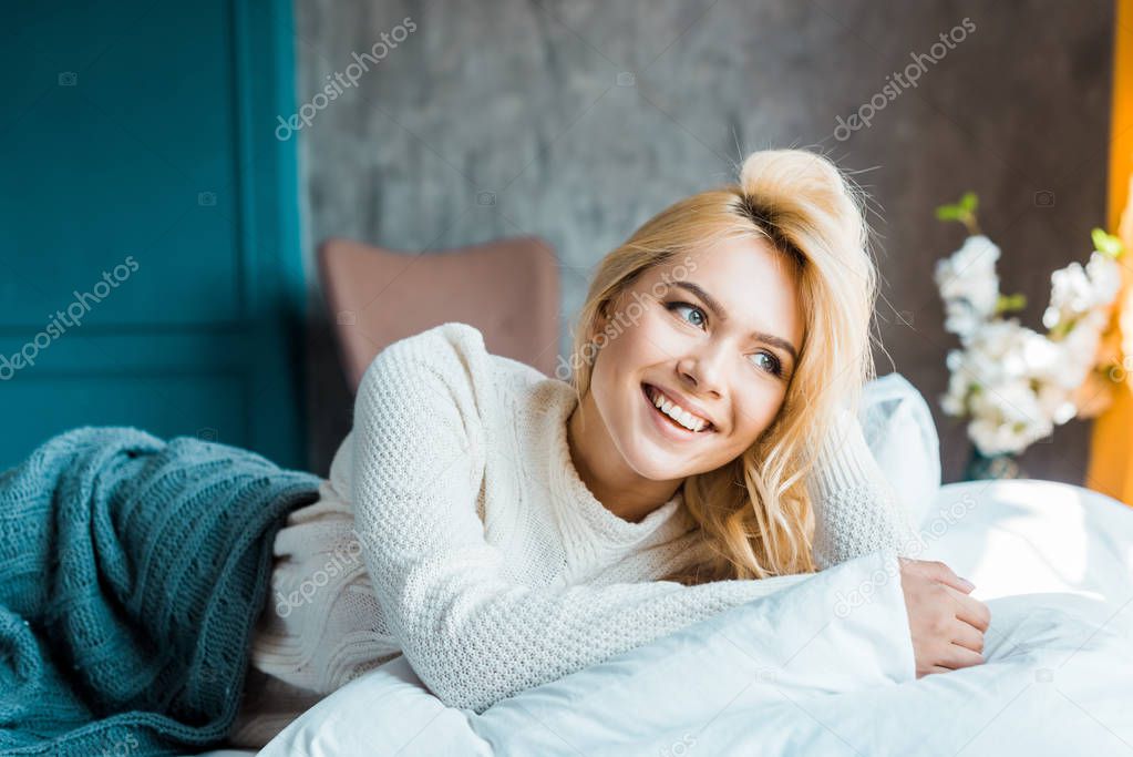 happy attractive woman in sweater lying under blanket on bed in bedroom and looking away