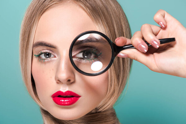 beautiful young woman holding magnifying glass and looking at camera isolated on blue