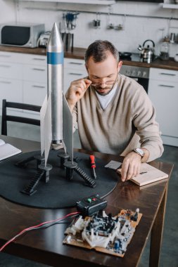 handsome engineer modeling rocket and looking above glasses at table at home clipart