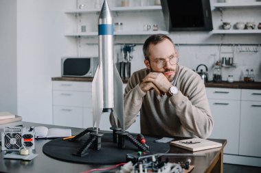 thoughtful engineer sitting at table with rocket model at home and looking away clipart