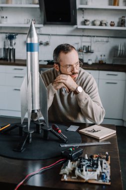 pensive engineer sitting at table with rocket model at home and looking away clipart