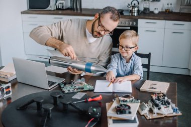 father and son testing rocket model together at home clipart