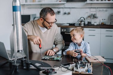 cheerful father and son repairing circuit board and looking at each other at home clipart