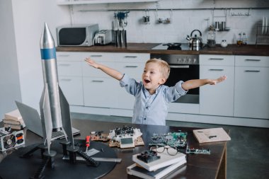 happy adorable boy standing with outstretched hands near rocket model in kitchen on weekend clipart