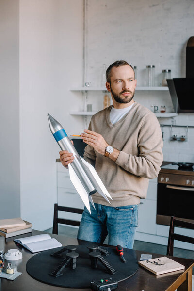 handsome engineer holding rocket model at home and looking away