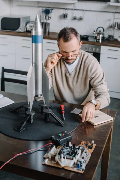 handsome engineer modeling rocket and looking above glasses at table at home