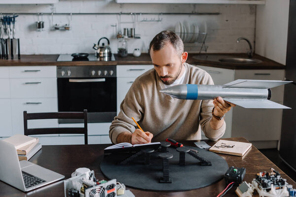 handsome engineer holding rocket model and making notes at home