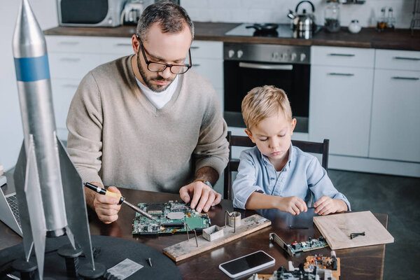 father and son soldering circuit board with soldering iron in kitchen