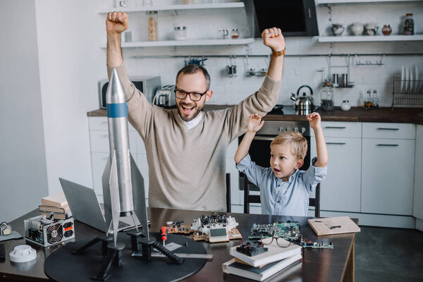 happy father and son with raised hands looking at rocket model at home