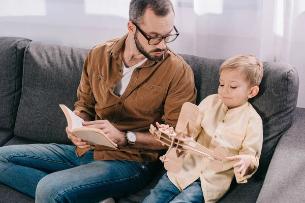 Father Holding Book Looking Son Playing Wooden Toy Plane Home — Free Stock Photo