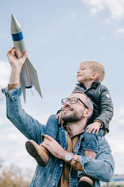 愉快的父亲背着小儿子颈部和钻孔模型火箭对天空 — 图库照片