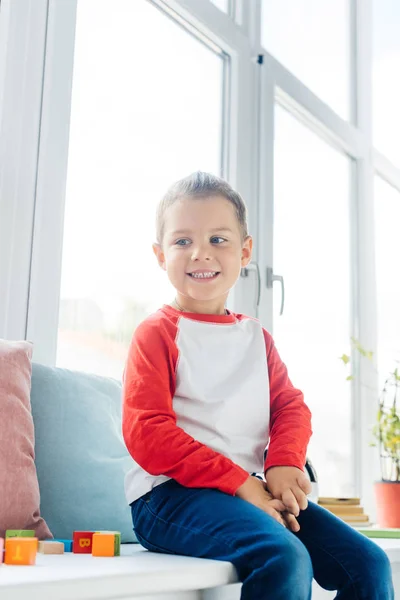 自宅の窓に座ってかわいい陽気な男の子の肖像画  — 無料ストックフォト