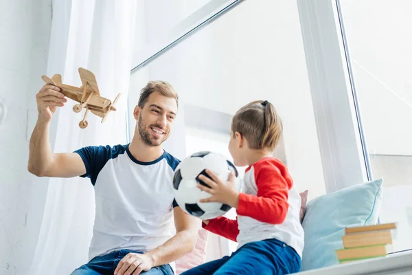 Junge Mit Fußballball Sieht Lächelnden Vater Mit Holzspielzeugflugzeug Hause — Stockfoto