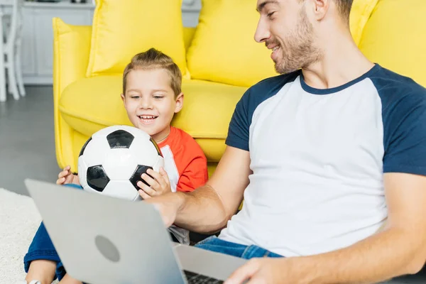 サッカー ボールの家の床に座って近くで幼い息子ながらノート パソコンを使用している人 — ストック写真