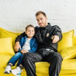 Handsome young father in police uniform watching tv with son while sitting on yellow couch at home