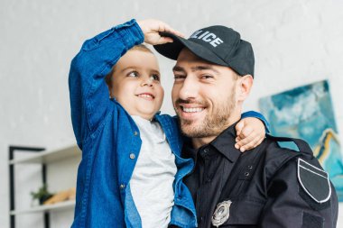 yakın çekim küçük oğlu taşıyan ve kameraya bakarak polis üniforması içinde gülümseyen genç baba portresi