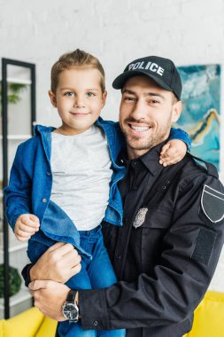 küçük oğlu taşıyan ve kameraya bakarak polis üniformalı genç baba gülümseyerek