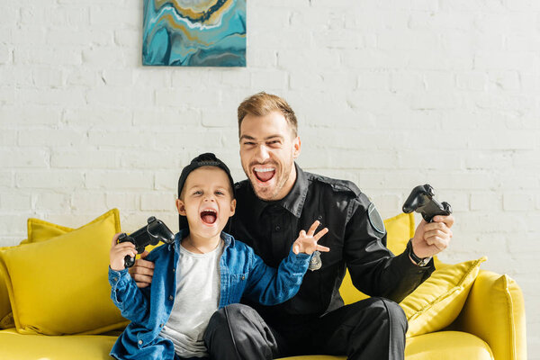 эмоциональный молодой отец в полицейской форме и сын играет в видеоигры, сидя дома на жёлтом диване
